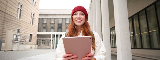 Happy redhead girl in red hat, walks around city with digital tablet, connects to public internet wifi and looks for route, looks at map on her gadget. - Foto, imagen