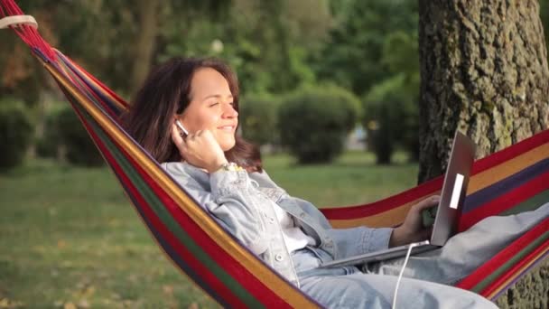 Hermosa mujer escuchando música en una hamaca en el parque usando un portátil. Recreación al aire libre. El trabajo de un freelancer. - Metraje, vídeo