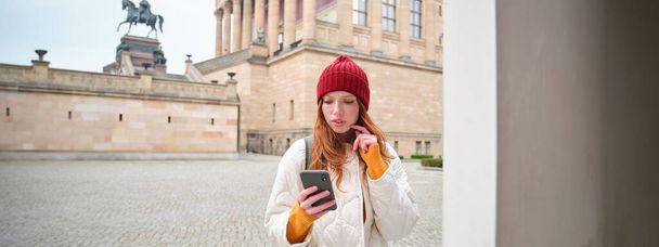 Smiling redhead girl tourist, walks around city and explores popular landmarks, sightseeing, holding smartphone, looking at her mobile app and checks with city map. - Photo, Image