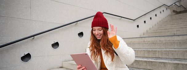 Радостная рыжая девушка подросток, студент с цифровым планшетом, говорит привет, волны рука на гаджет-камеру, подключается к видео-чату, разговаривает с другом в приложении. - Фото, изображение