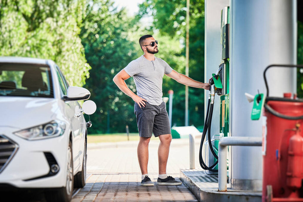 Driver with gasoline pump refilling car gas tank. Confident man refueling his luxury white auto. Man in casual clothes and sunglasses at modern gas station. - Photo, Image