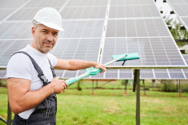 Muž čistí solární fotovoltaický panel pomocí mop. Profesionální pracovník odvádí dobrou práci při čištění solárního panelu od prachu. Muž držící mop a usmívající se přímo do kamery. - Fotografie, Obrázek