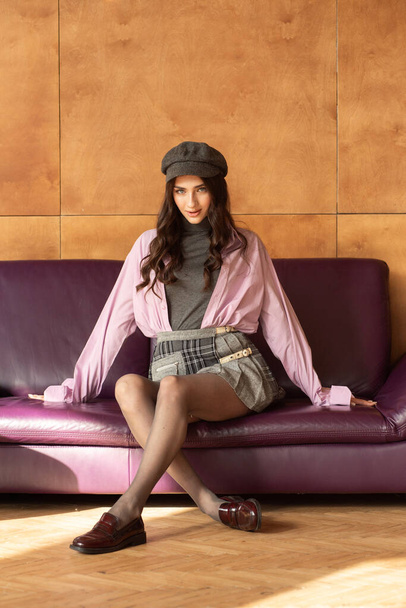 beautiful young woman with stylish hair in a hat and a leather jacket sitting on the couch - Photo, Image