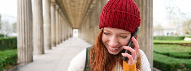 Portrait of redhead european girl in red hat, makes a phone call, walks in city and talks to friend on smartphone. Copy space - Photo, image