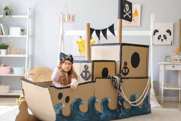 Schattig klein meisje verkleed als piraat spelen met spiegelglas in karton schip thuis - Foto, afbeelding