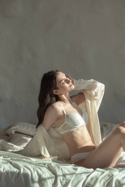 mladá krásná brunetka modelka pózující ve studiu, sedí v bílé posteli - Fotografie, Obrázek