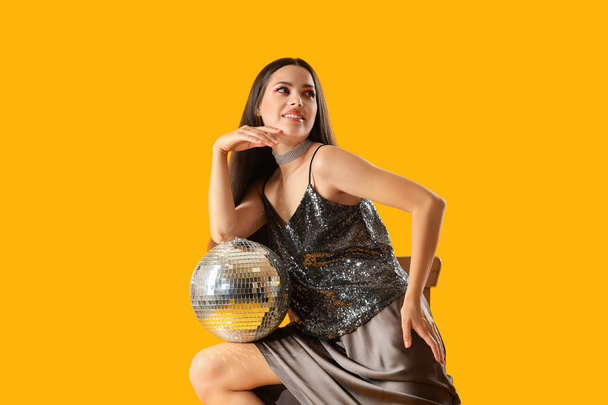 Stylish young woman with disco ball sitting on chair against orange background - Foto, Imagen