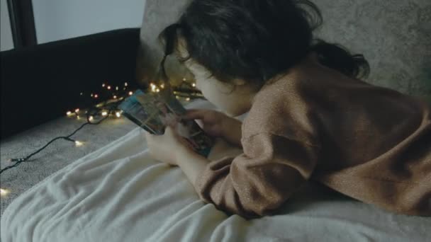 Egy kislány régi képeket néz egy karácsony estéjén. Karácsonyi fények körül. Karácsony és szabadidő koncepció - Felvétel, videó