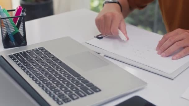 Close-up van handen van onherkenbare man die werkt of studeert op laptop en aantekeningen maakt in copybook from home - Video