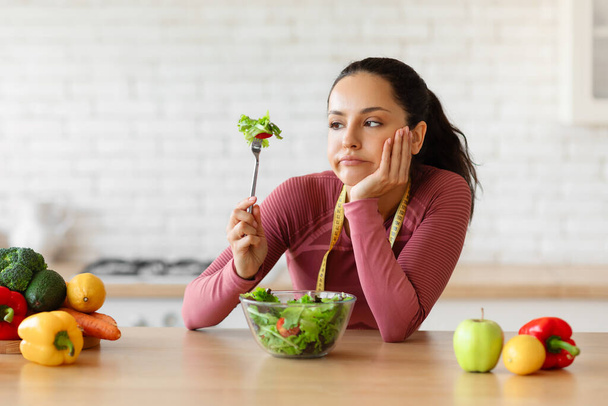 ダイエットの戦い。 野菜サラダを食べるアクティブウェアのフラストレートフィット感の女性と怒りの表情,ダイエットメニューの疲れ,家庭での現代キッチンでの食欲の欠如を見て - 写真・画像
