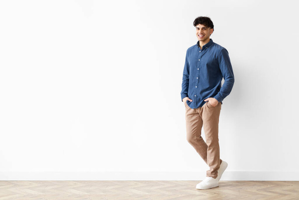 Confident Person. Full length shot of cheerful casual arab man posing standing isolated over white background, leaning on wall holding hands in pockets, smiling to camera. Copy space - Photo, Image