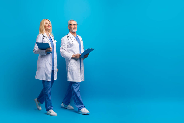 Επαγγελματική ιατρική ομάδα δύο ηλικιωμένοι γιατροί άνδρες και γυναίκες που κατέχουν ιατρικά διαγράμματα έχουν ιατρικό consilium, τα πόδια προς το χώρο αντιγραφής και χαμογελώντας, μπλε φόντο στούντιο, πλήρους μήκους. Σύγχρονη υγειονομική περίθαλψη - Φωτογραφία, εικόνα