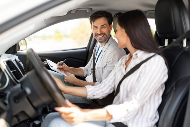 Przystojny instruktor mężczyzna badający szczęśliwą studentkę, robiąc notatki na wykresie, siedząc przy wesołej młodej kobiecie prowadzącej auto, widok z boku, przestrzeń do kopiowania. Koncepcja szkoły jazdy - Zdjęcie, obraz
