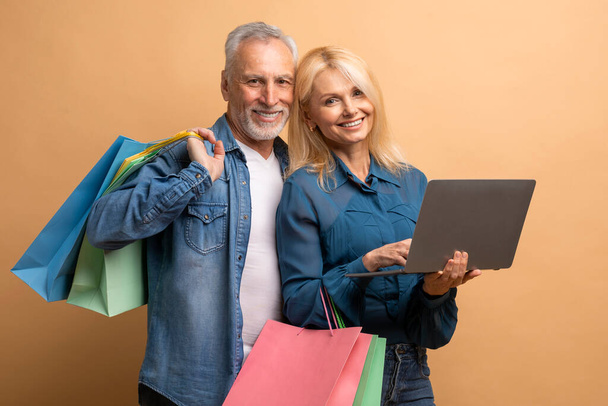Cheerful attractive caucasian elderly man and woman shopping on Internet together, using laptop computer pc, holding purchases, beige background. Retail, ecommerce - Photo, Image
