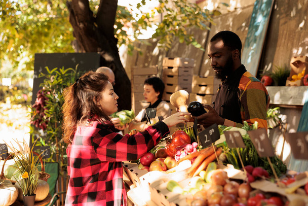 Afrikkalainen amerikkalainen kaveri myyjä seisoo takana pilttuu myyvät tuoreita orgaanisia kauden vihanneksia naispuolinen asiakas paikallisten viljelijöiden markkinoilla. Nuori nainen ostaa luonnollisia kotimaisia tuotteita - Valokuva, kuva