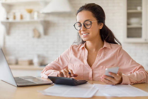 Šťastná žena s brýlemi sedí u kuchyňského stolu a komunikuje se svým notebookem, telefonem a kalkulačkou. Dokumenty jsou rozloženy, označují práci nebo finanční sezení - Fotografie, Obrázek