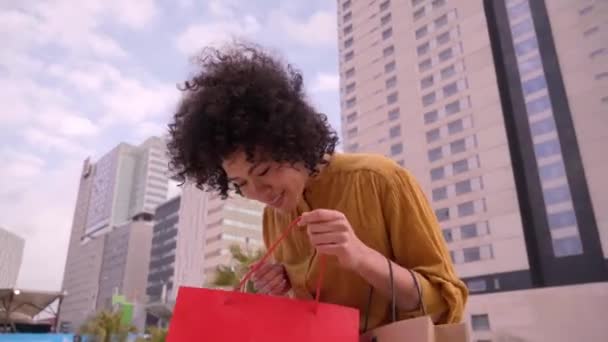 Video of surprised young African American female shopaholic holding paper bags with shopping and looking at camera standing dancing outside mall. People excited on black Friday. Sales season. - Footage, Video