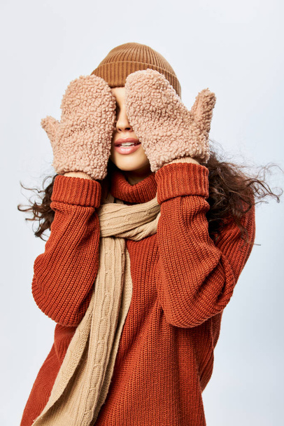 young woman in hat and knitted terracotta sweater covering eyes with mittens on grey backdrop - Photo, Image