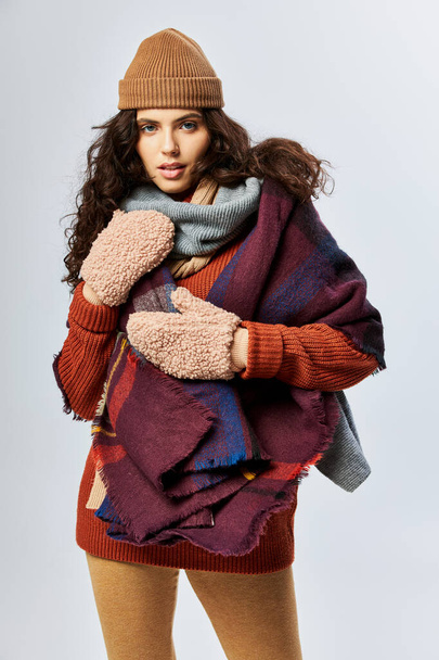 ζεστά στρώματα, σγουρά νεαρή γυναίκα σε στρώματα ρούχα με ζεστό κασκόλ και γάντια σε γκρι φόντο - Φωτογραφία, εικόνα