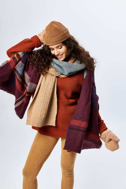 Kış tarzı, katmanlı giysiler içinde neşeli genç bir kadın, gri arka planda örülü şapka ve eldivenler. - Fotoğraf, Görsel