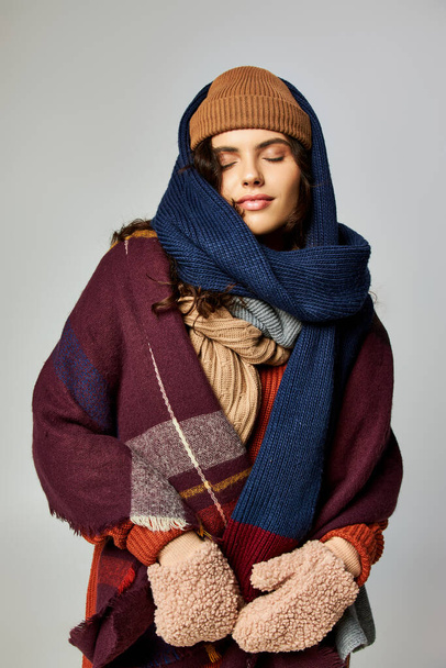 estilo invierno, mujer joven en capas de ropa, sombrero de punto y bufandas posando sobre fondo gris - Foto, imagen