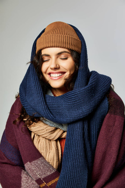 winter mode, vrolijke vrouw in gelaagde kleding, gebreide muts en sjaals poserend op grijze achtergrond - Foto, afbeelding