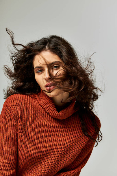 wind blowing at face of brunette curly woman in terracotta sweater on grey backdrop, winter fashion - Photo, Image