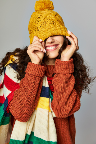 ευτυχισμένη γυναίκα φορώντας ζεστό καπέλο bobble και πουλόβερ με γυμνό κασκόλ ποζάρουν σε γκρι φόντο - Φωτογραφία, εικόνα