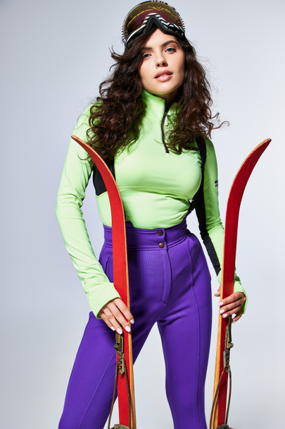 attractive young woman with curly hair posing in trendy active wear and holding skis on grey - Photo, Image