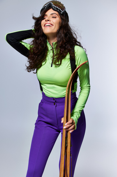 joyeuse jeune femme aux cheveux bouclés posant en tenue active tendance et tenant des skis sur gris - Photo, image