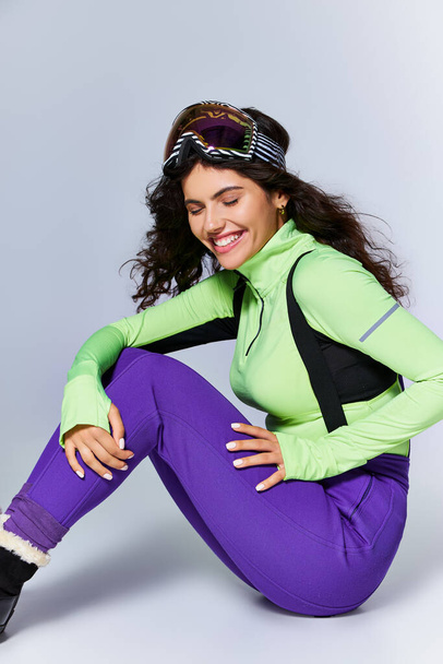 zimní sport, veselá a sportovní žena s kudrnatými vlasy sedí v aktivním oblečení na šedém pozadí - Fotografie, Obrázek
