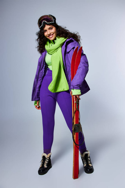 Wintersport, fröhliche Frau mit lockigem Haar posiert in aktiver Kleidung mit Kugeljacke und Skiern - Foto, Bild