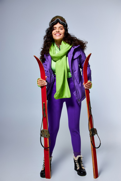 winter sport, positive woman with curly hair posing in active wear with puffer jacket and skis - Photo, Image