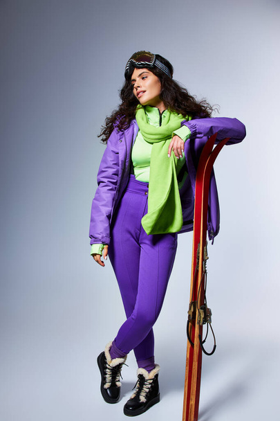 Winteraktivität, charmante Frau mit lockigem Haar posiert in aktiver Kleidung mit Kugeljacke und Skiern - Foto, Bild