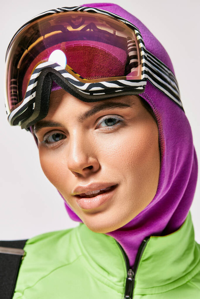 mujer con estilo en ropa de esquí de invierno con pasamontañas en la cabeza mirando a la cámara en el fondo gris - Foto, Imagen