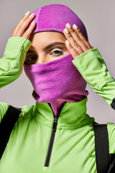 portrait of woman with blue eyes wearing purple ski mask and looking at camera on grey background - Photo, Image