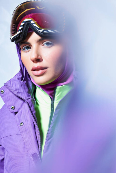 привлекательная женщина с голубыми глазами, позирующая в лыжных гуглях, балаклаве и зимней куртке на сером фоне - Фото, изображение