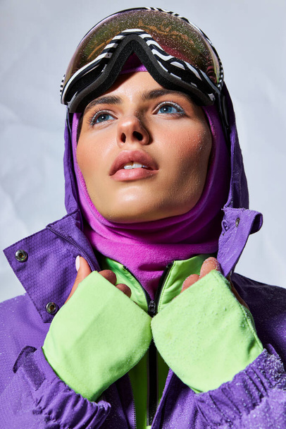temps froid, femme rêveuse en cagoule et googles de ski posant en veste d'hiver pourpre sur gris - Photo, image