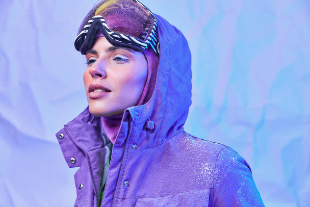 fagyos téli levegő, fiatal nő símaszkban, googles és meleg kabát néz félre lila háttér - Fotó, kép
