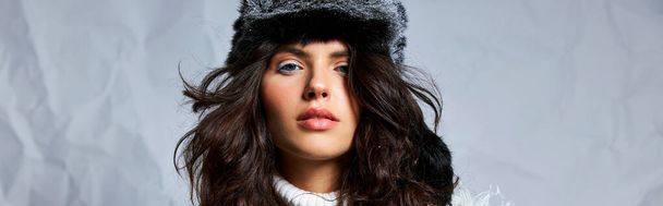 göndör nő műszőrme kalap és pulóver nézi kamera szürke háttér, téli szépség transzparens - Fotó, kép