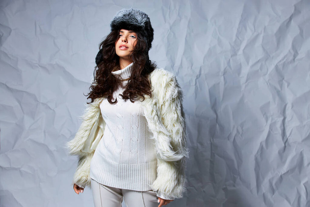 gorgeous curly woman in white faux fur jacket, winter hat and sweater posing on grey backdrop - Foto, Imagem