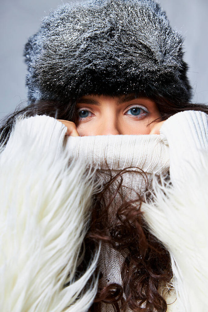 stunning woman in white faux fur jacket, winter hat and sweater posing on grey backdrop, blue eyes - Photo, Image