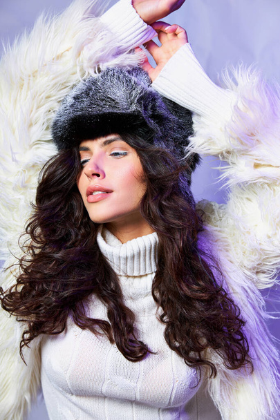 curly brunette woman in faux fur jacket, winter hat and sweater posing with raised hand on blue - Photo, Image