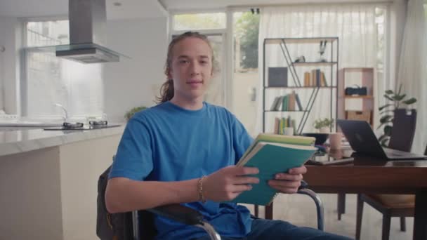 Keskipitkä muotokuva nuoresta positiivisesta valkoihoisesta opiskelijasta pyörätuolissa pitäen hallussaan oppimateriaalia ja katsoen kameraa kotona - Materiaali, video