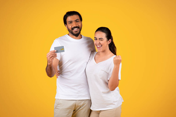 Glad millennial arabic husband and wife show credit card, rise fist, celebrate win, success, isolated on yellow studio background. Online shopping, credit, recommends sale, profit and savings - Photo, Image