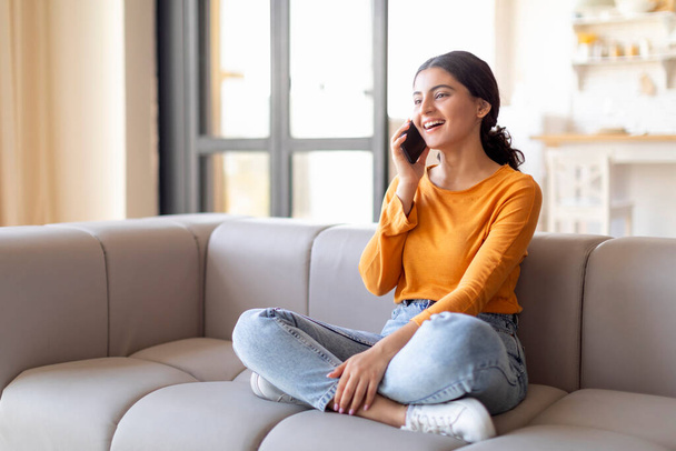 Portrait Of Beautiful Young Indian Woman Talking On Mobile Phone At Home, Smiling Millennial Eastern Female Enjoying Pleasant Cellphone Conversation, Sitting On Couch In Living Room Interior - Photo, Image