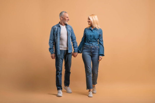Loving happy senior couple holding hands and walking together isolated on beige studio background, looking at each other, have conversation. Elderly man and woman have romantic date, full length - Valokuva, kuva