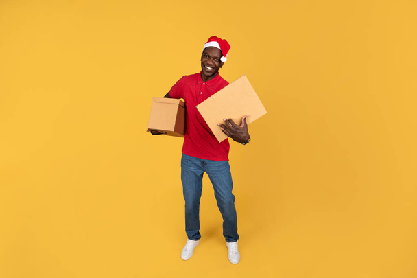 Cheerful young black delivery man in Santa Claus hat hold cardboard boxes, has fun, isolated on orange studio background. Order delivery service, work for Xmas, New Year - Photo, Image