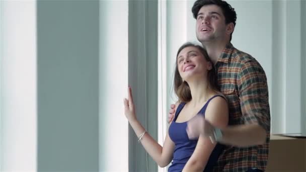 Μετακίνηση σε μια νέα ζωή. Ένα κορίτσι και έναν άντρα που κατέχουν τα κουτιά για την κίνηση τα χέρια και να χαμογελά στη φωτογραφική μηχανή, ενώ ένα ζευγάρι ερωτευμένο στέκεται στο παράθυρο μεταξύ κουτιά - Πλάνα, βίντεο