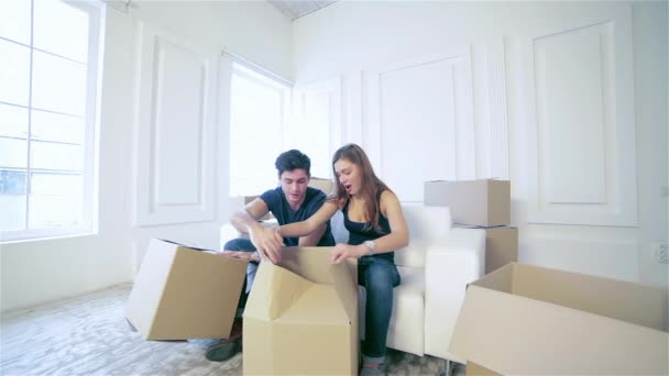 Shifting to new life. Couple in love moving and keep a box in his hands and looking inside box while a young and beautiful couple in love sitting on the couch in an empty apartment among boxes - Footage, Video
